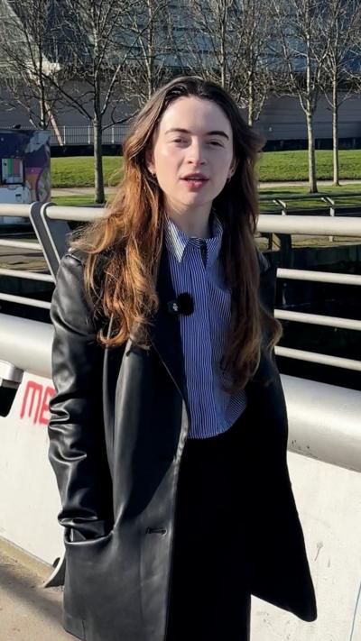 A young woman with brown hair walks on a bridge that has grey railings. It is a sunny day and she wears a blue striped shirt and black leather coat.