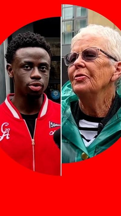 A young man in a red jacket and an elderly woman in a green coat speak on a street
