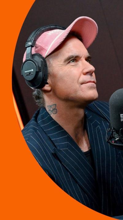 A man, Robbie Williams, in the BBC Radio 2 studio wearing a pink cap and pin striped