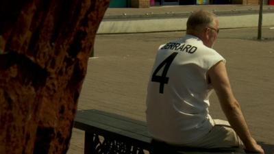 Man in Gerrard England football shirt sitting on bench