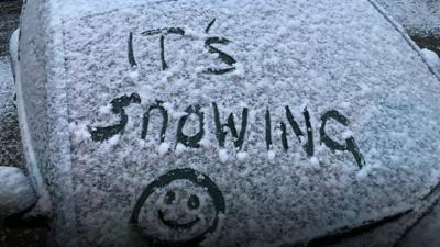 A light dusting of snow on a car with the words "it's snowing" written into the snow.