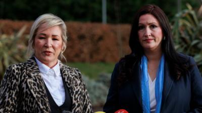 Michell O'Neill and Emma Little Pengelly standing next to each other wearing coats. They are outside and there is some grass and hedges in the background.