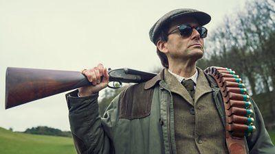 David Tennant in a green coat, and a flat cap, wearing sunglasses with a shotgun over one shoulder and ammo over the other