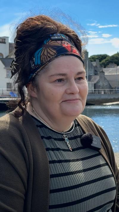 Diane Barnes has dark hair, she is sitting in a harbour area, wearing a green top, necklace and a bandana.