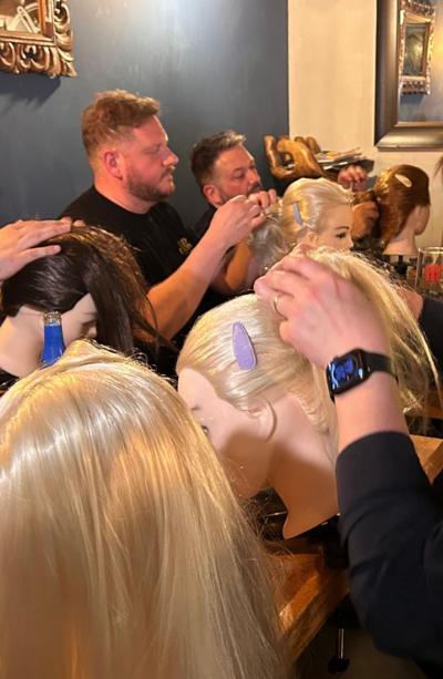 Dads sat around a table, each have a mannequin head in front of them and hair. They're being taught how to plait hair and have their hands in the hair to style.