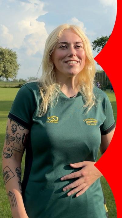A woman with shoulder-length blonde hair smiles at the camera. She wears a green sports t-shirt and her right arm is covered in patchwork-style tattoos.