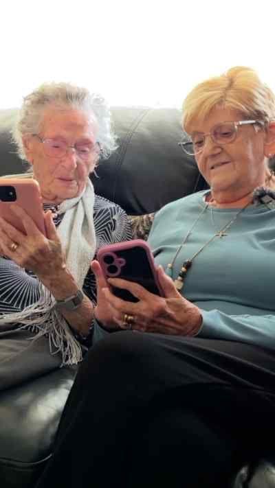 Two women sit next to each other on a sofa. They are both looking at their mobile phones.