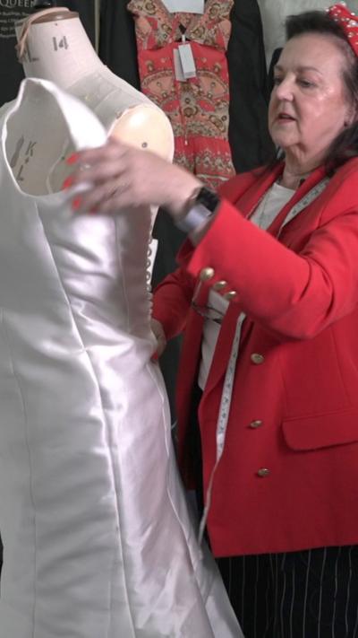 A lady in her sixties, wearing a red jacket and white top, with a measuring tape around her shoulders, works on a wedding dress on a mannequin.
