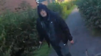 A man in a black hooded top and a tracksuit. He is standing on a pavement with bushes either side of him, in a residential area. He is holding a large knife in his right hand. His face is blurred to obscure his identity.