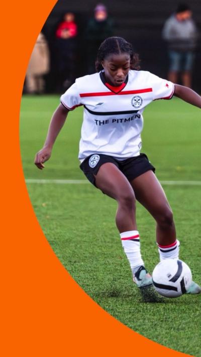 A young woman in a football kit controls the ball with her feet while on the pitch