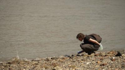 Woman bends down to inspect something on shore of the Thames