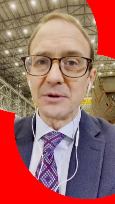 Chris Mason stands in a hall - he has short brown hair, and wears brown glasses and a dark blue suit jacket with light blue shirt and patterned tie