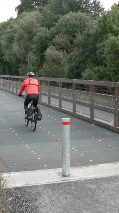 A man on a bicycle, cycling along a road. There are trees to the right of the road alongside a fence. The man is wearing a red jacket, black trousers and a black helmet. He is pictured from behind.