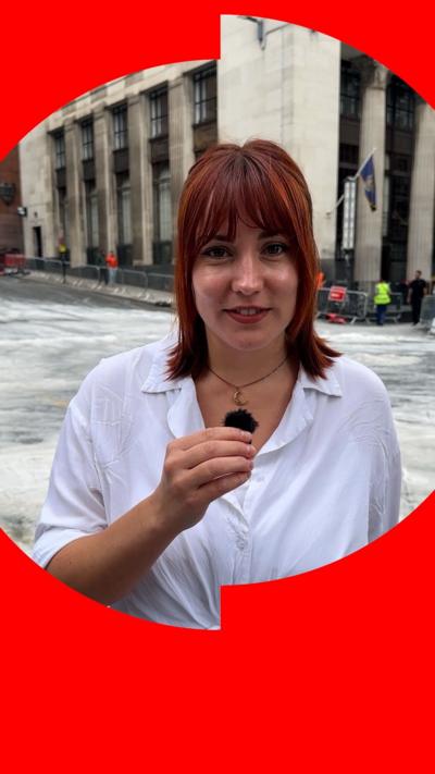 A woman with red hair stands in the middle of a Glasgow street. She is wearing a white shirt and holding a small mic in front of her.