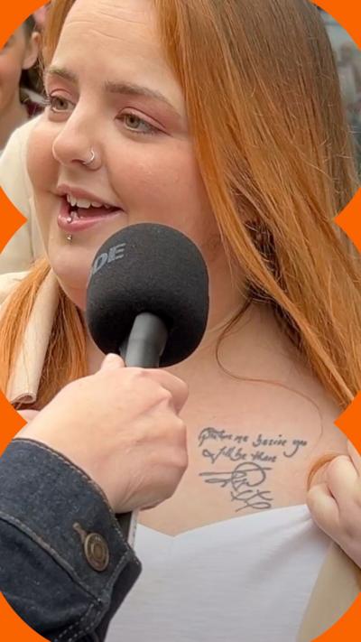 A woman with dyed ginger hair pulls her top to one side to show a tattoo below her collar bone