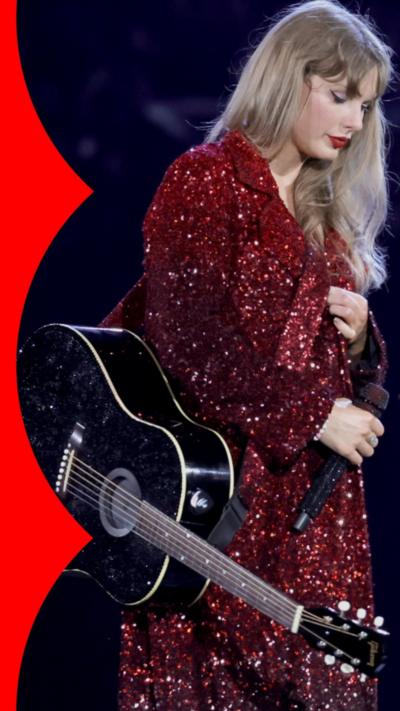Taylor Swift is wearing a red sparkly dress and stands with a guitar hanging to her right hand side.