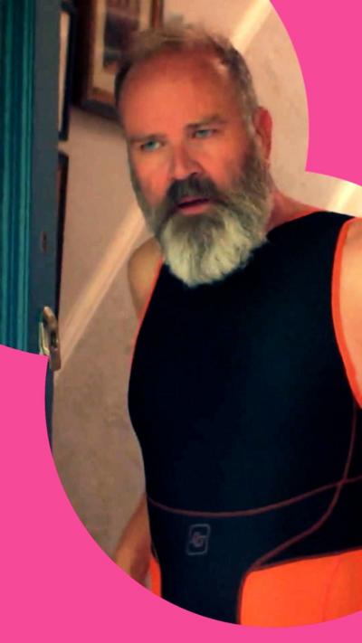 An older man with grey hair and beard wears a sleeveless, black and orange sporting catsuit. He is standing in a front doorway.