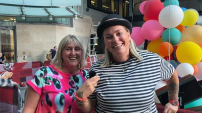 Two smiling women, Take That fans, stand outside Broadcasting House in central London. One is blonde and is wearing a neon pink t-shirt with some blue and black splotches. The other is blonde and wearing a bowler hat and a stripy black and white t-shirt. 