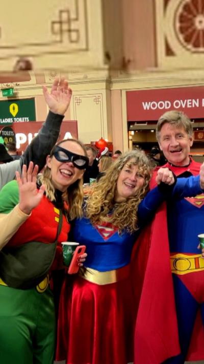 Two women and a man wearing superhero outfits holding drinks and cheering