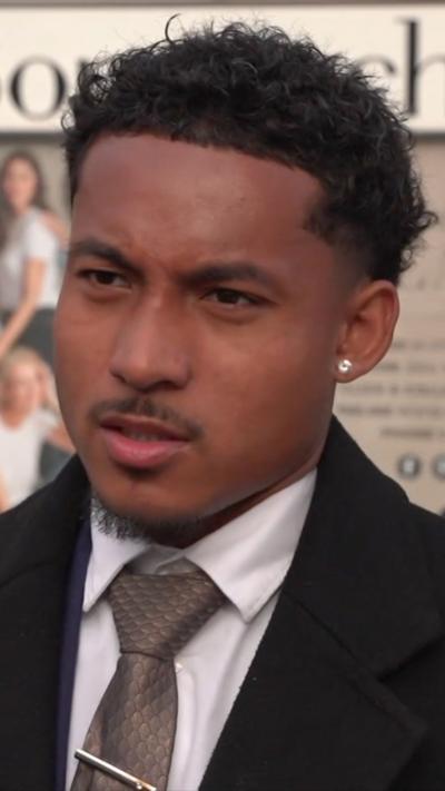 A man with short curly hair looks off camera with a furrowed brow. He has a moustache, some facial hair on his chin, dark curly hair on his head and an ear stud. He is wearing a white shirt buttoned up with a tie and a black jacket.