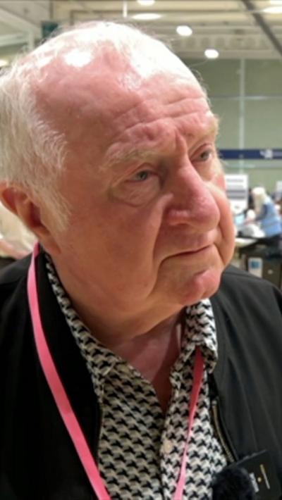 Grey haired man with black jacket and white and black patterned shirt, with a red lanyard