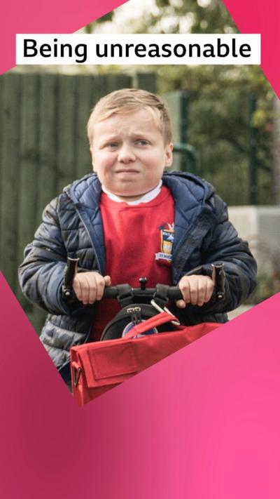 A boy, the character Ollie, grimaces - he wears a red school jumper and blue coat