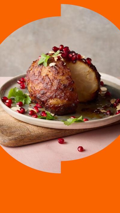A golden roasted celeriac sits on a round plate. It is garnished with pomegranate seeds, coriander and flaked almonds