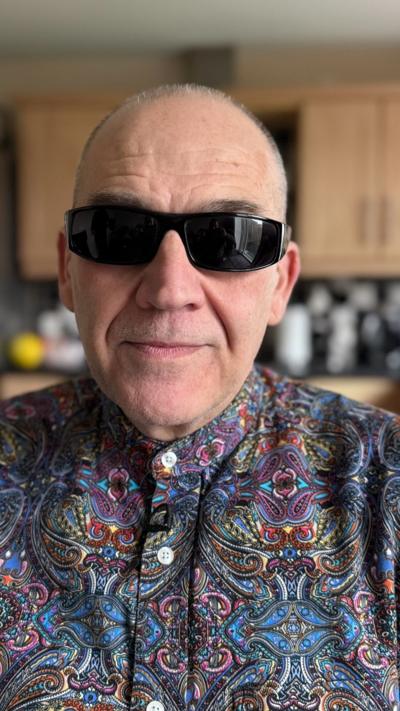 A man with dark glasses and floral shirt looking at the camera