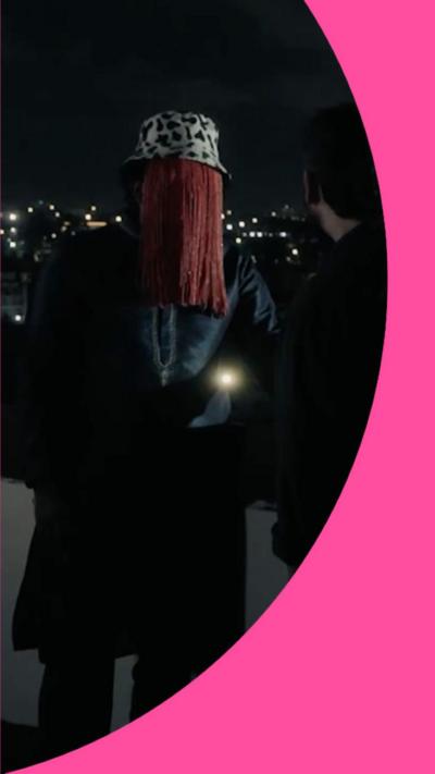 A man wearing a Dalmatian print hat with red tassels hanging from the edges to cover his face stands on a rooftop next to another man. Both are dressed in black.