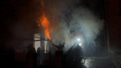A figure holds an exploding firework in their hand, with smoke and other hooded figures crowded around outside a house. 