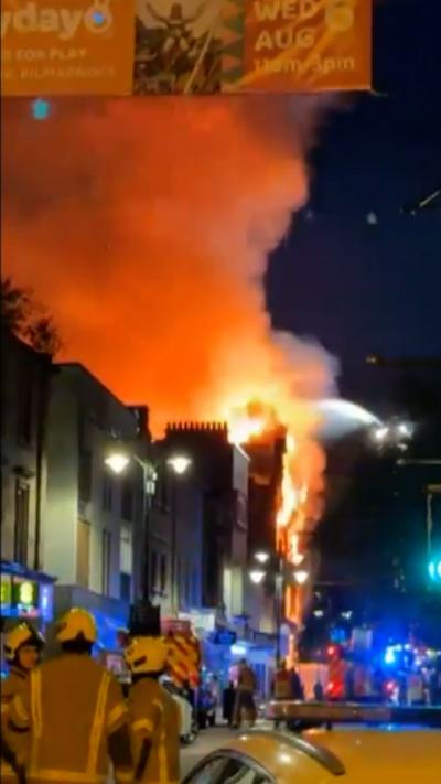 Firefighters look at flames and smoke coming out the top of a building