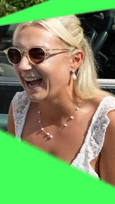 A blonde woman dressed in white, with sunglasses, pearl earrings and necklace laughs on a sunny day.