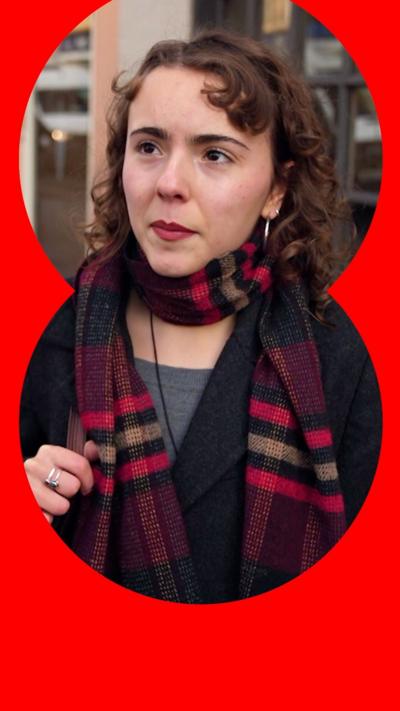 A woman with curly brown hair is interviewed in the street. She wears a maroon checked scarf and black coat