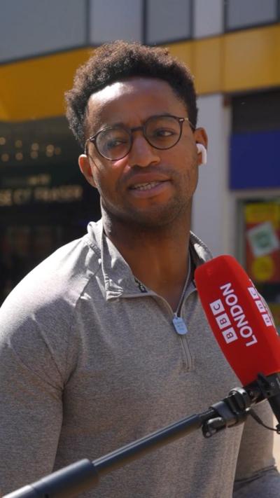 Person wearing a grey zip-up shirt and AirPods speaking into a BBC London microphone outdoors near storefronts.