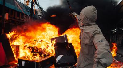 Hooded man brandishes a black stick towards a burning building