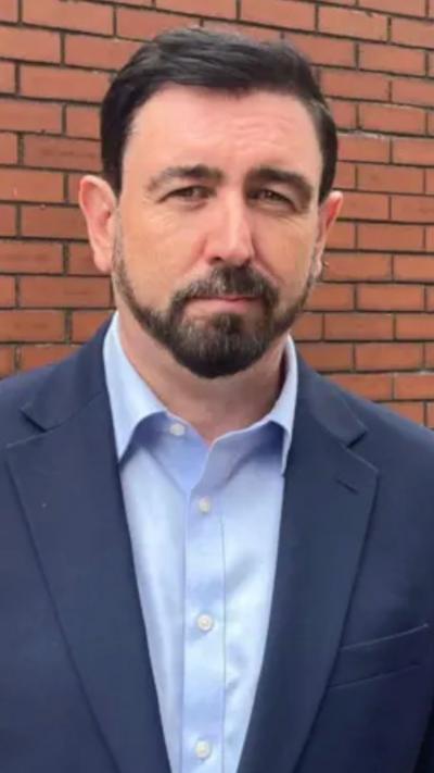 man in a suit looking seriously at the camera, in front of a red brick wall