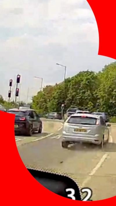 Screenshot of dashcam footage showing a silver car on a road, with a roundabout ahead