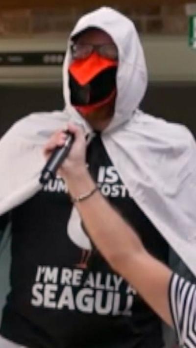 Seagull impersonator with white cloak and orange beak. He is speaking into a microphone held by another person.
