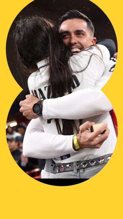 A man and a woman embrace. He is smiling and holding a small box. She is facing away from the camera