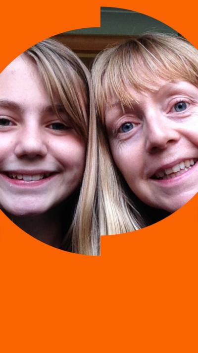 A young girl smiles in a selfie with her mother, in front of kitchen cupboards. Both have blonde hair.