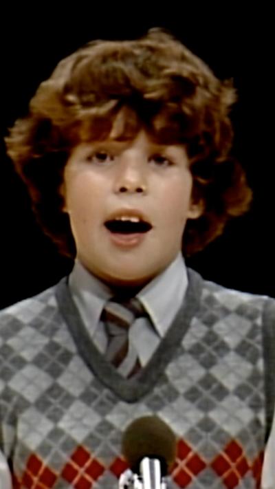 Rhun ap Iorwerth, the first minister of Wales, is pictured singing in the Urdd Eisteddfod in 1983, he is a child with thick long brown hair and a white collared shirt and grey sweater. The background is black.
