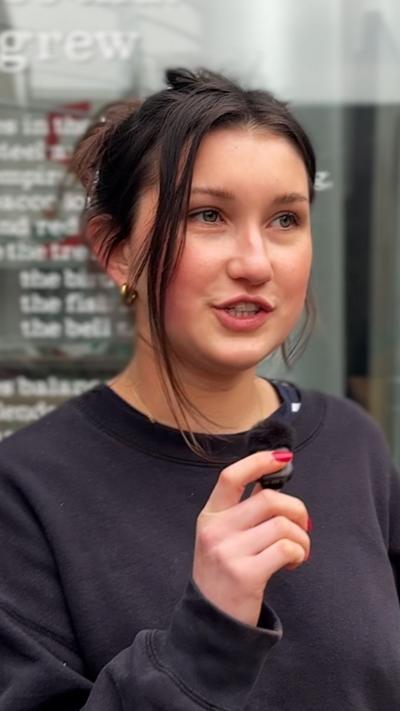 A girl with her mouth slightly open looks off to the side. She has dark hair that is tied up and hazel eyes. She is wearing gold earrings and a dark jumper. She stands in front of a glass window that has text on it.