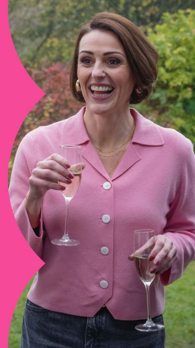 Character Suz laughs in a garden while holding a glass of fizz. She has brown hair and wears a pink shirt