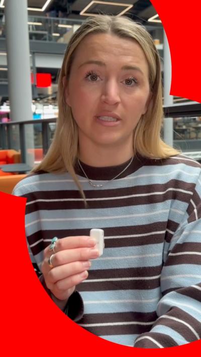 Lucy Woodham looks at the camera while holding a small white mic. She is wearing a blue and brown striped jumper.