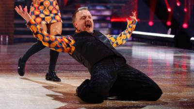 Chris McCausland, wearing a brightly coloured orange shirt and black waistcoat, slides on the dancefloor on his knees with his arms in the air and his mouth open.