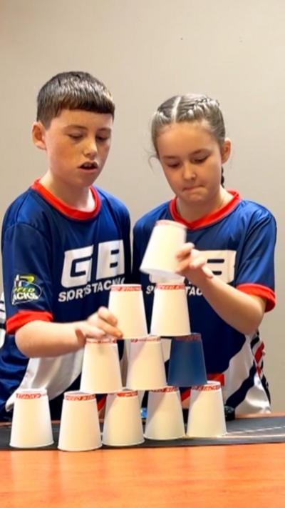 Two children - a boy with short brown hair and a girl with blonde hair pulled back behind her head - are stacking white cups in a pyramid. They are wearing dark blue sports tops with "GB sport stacking" written on them in white.