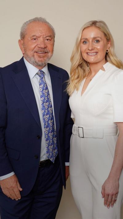 Sir Alan Sugar stands to the left of the image smiling. He is dressed in a dark blue suit. Rachel Woolford stands next to him, she wears a white jumpsuit with a belt at the waist