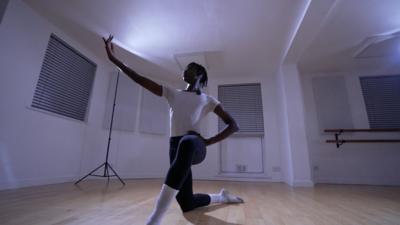 A young ballet dancer wears white socks, a t-shirt and tights. One hand is on his hip and the other is poised straight out as he kneels on the floor. 