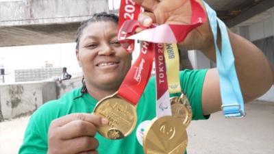 Foleshade Oluwafemiayo in green t-shirt holds up three gold medals