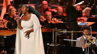 Samara Joy, wearing white and singing into a microphone, on stage at the Proms with musicians around her.
